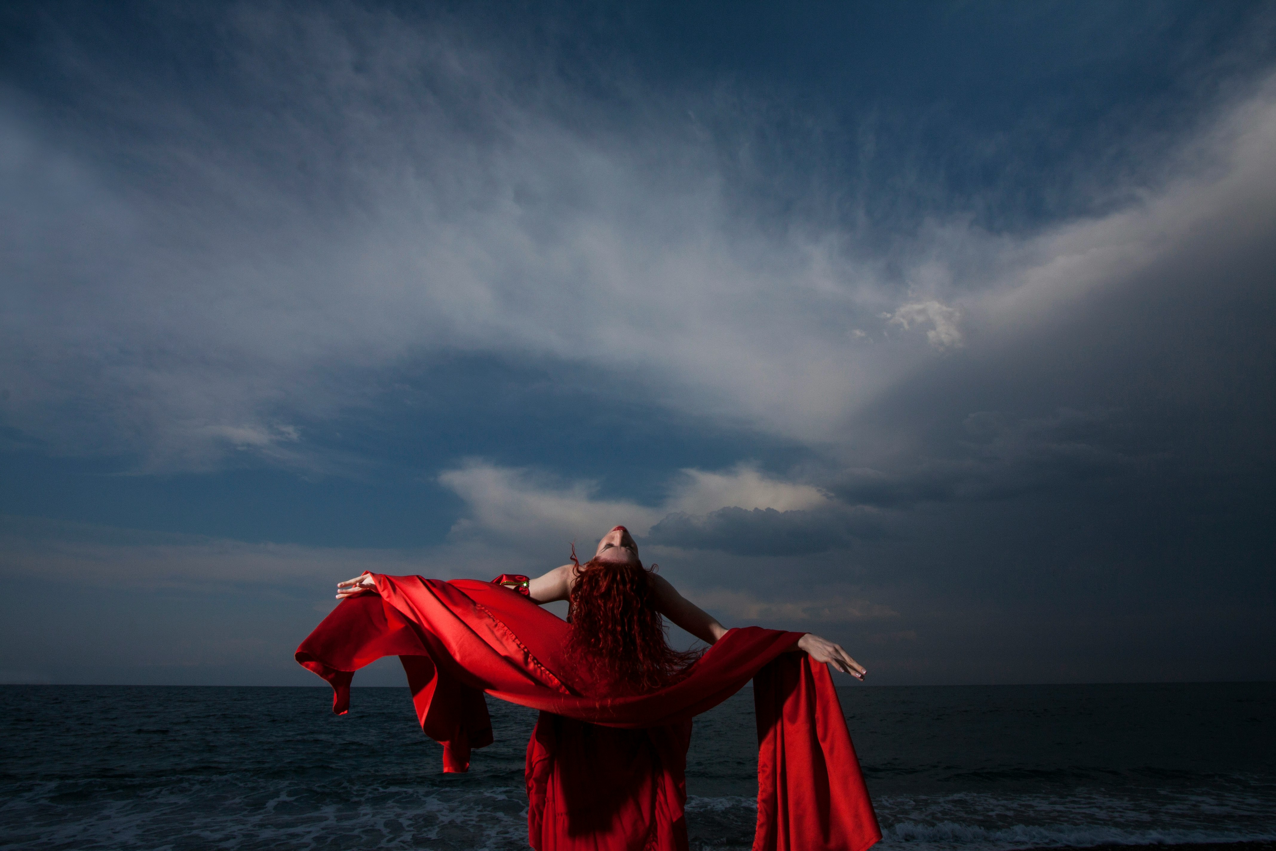woman in red dress standing on sea shore under gray cloudy sky during daytime